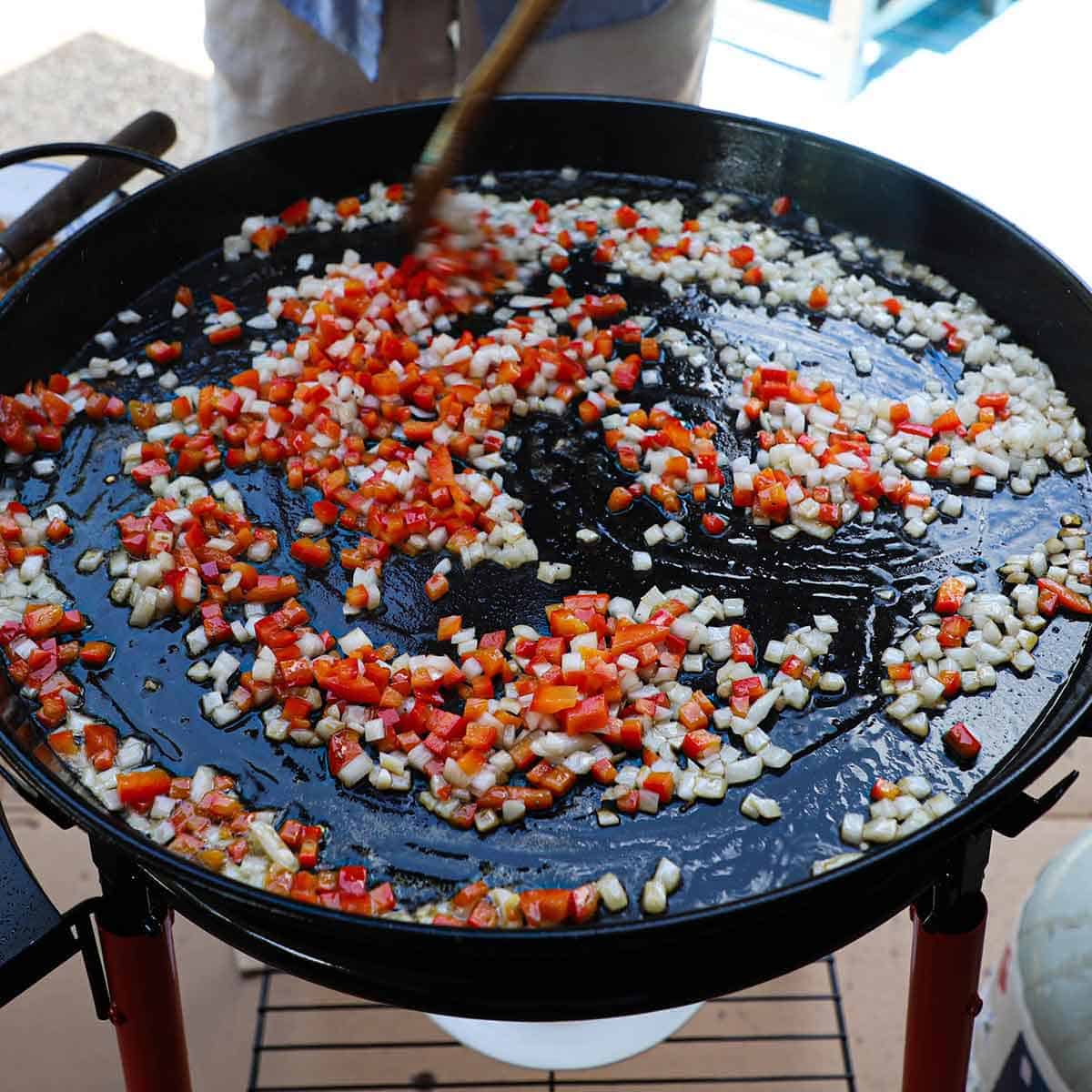 A person using a wooden spatula to stir chopped onion and red bell pepper in hot oil in a large paella pan.