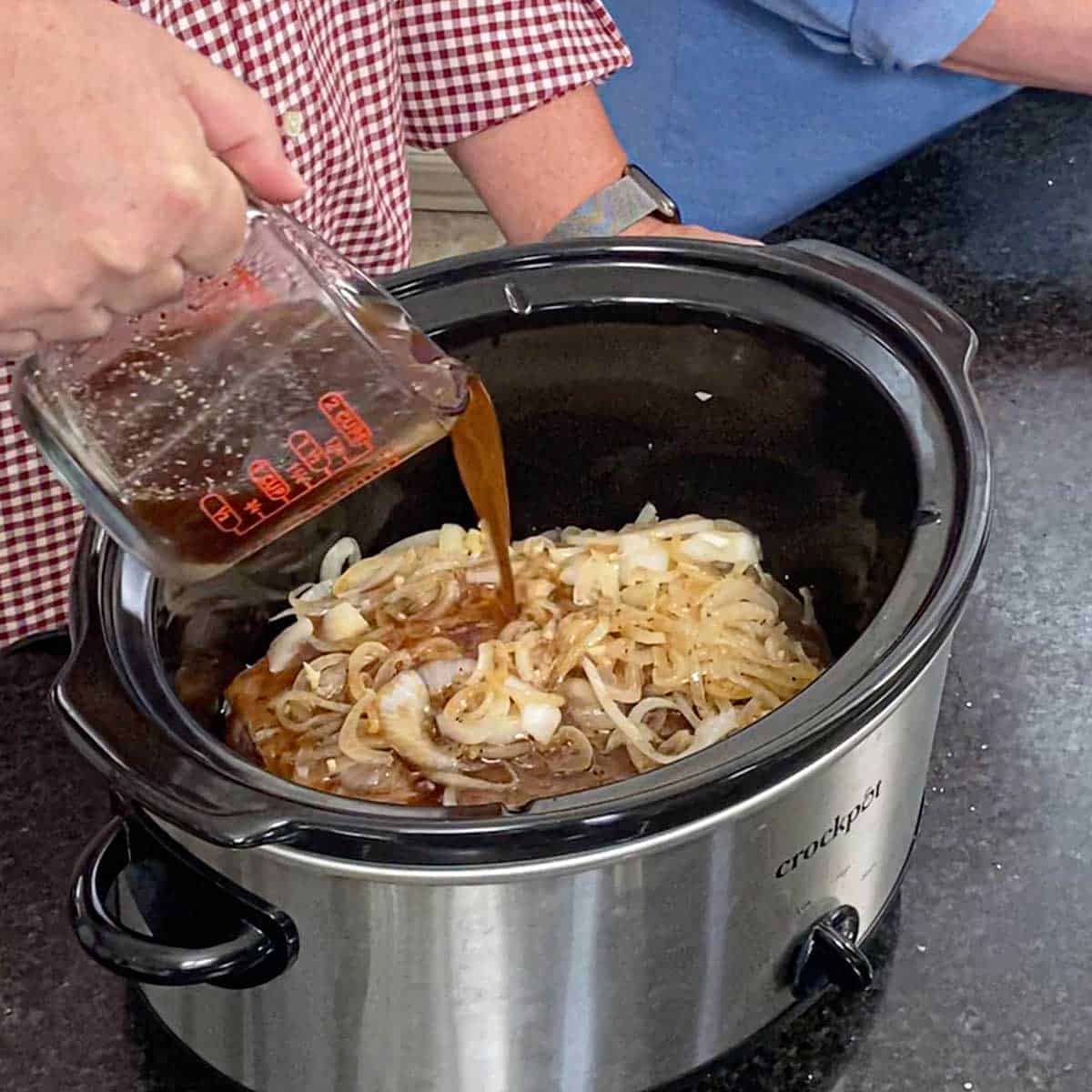 A person pouring beef stock from a glass measuring cup into a slow cooker that contains a partially seared chuck roast that is topped with strips of sautéed onions.