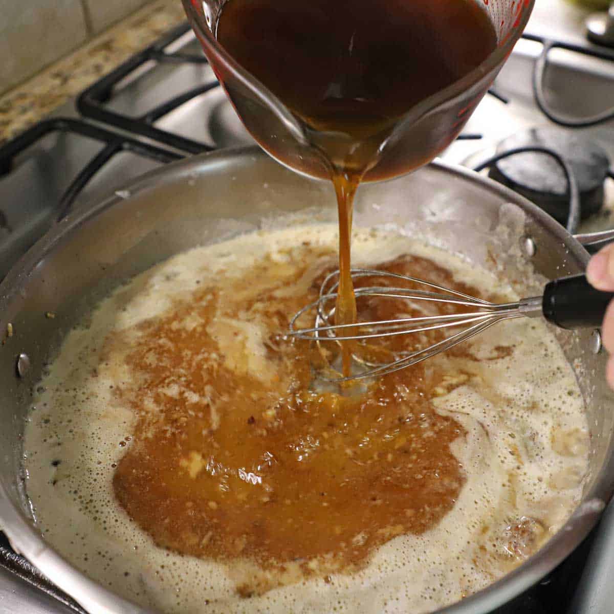 A person pouring beef stock into a skillet filled with a simmering roux that is being whisked by his other hand.