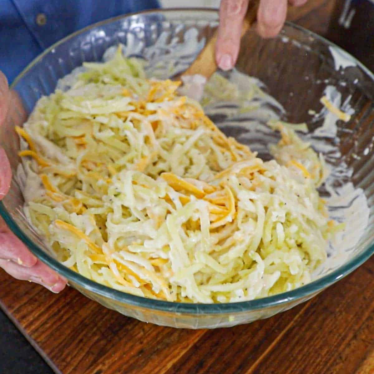 A person using a large wooden spoon to stir shredded potatoes, shredded cheddar cheese, chopped onions, and a homemade cream of chicken sour cream sauce in a large glass bowl.