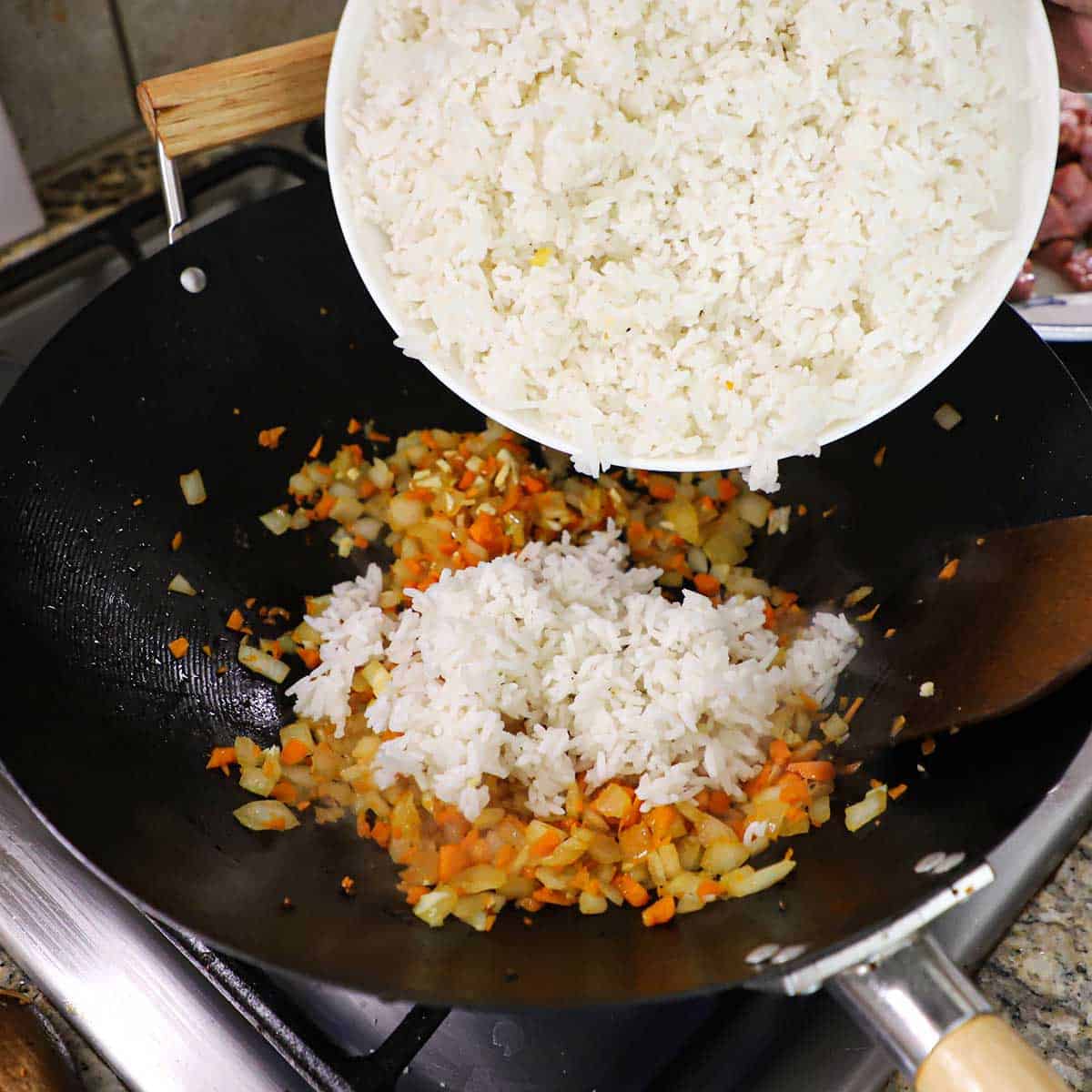 A person transferring a bowl of day-old white rice into a large wok filled with carrots, onions, garlic, and ginger that are being stir-fried.