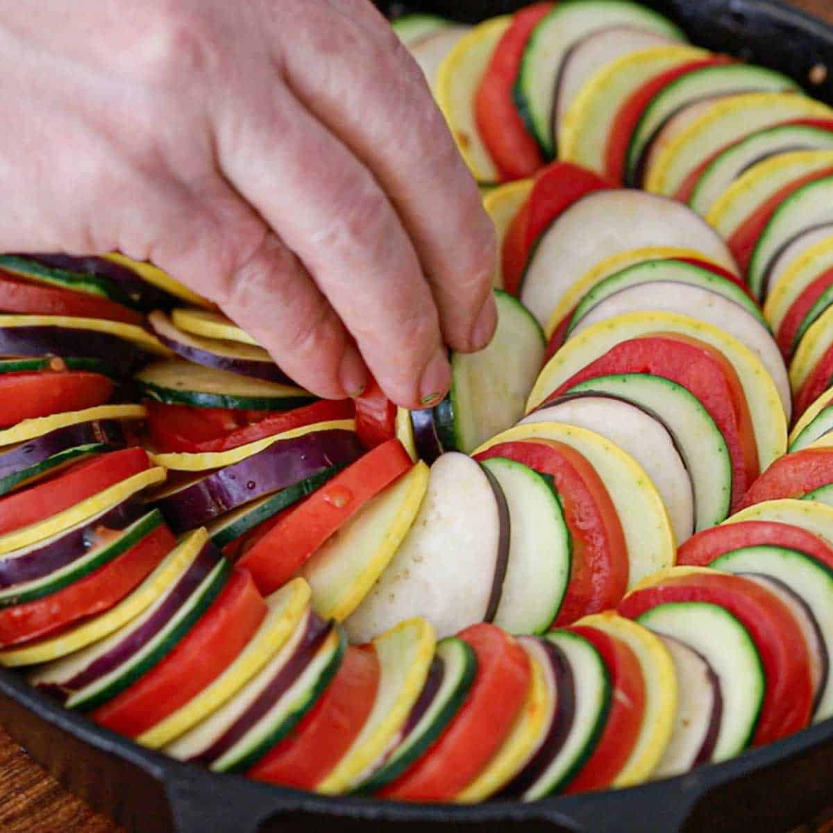A person placing a small stack of sliced vegetables on their side in the middle of an uncooked cast-iron skillet ratatouille.