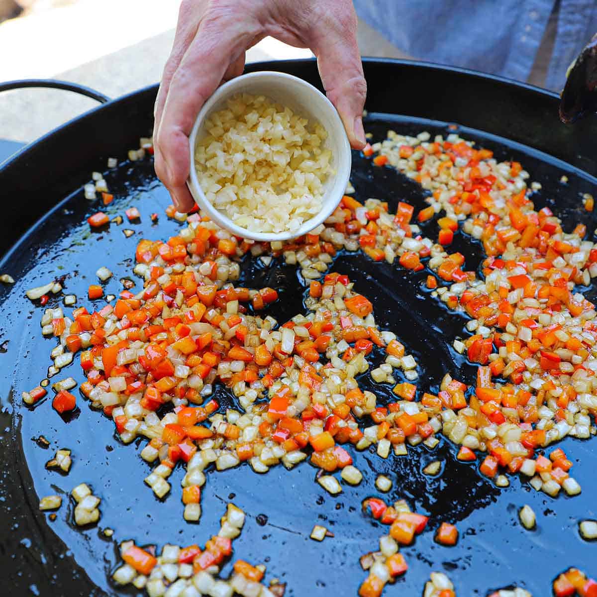 A person transferring minced garlic into a large paella pan that contains chopped onions and red bell pepper that are being sautéed in hot oil.