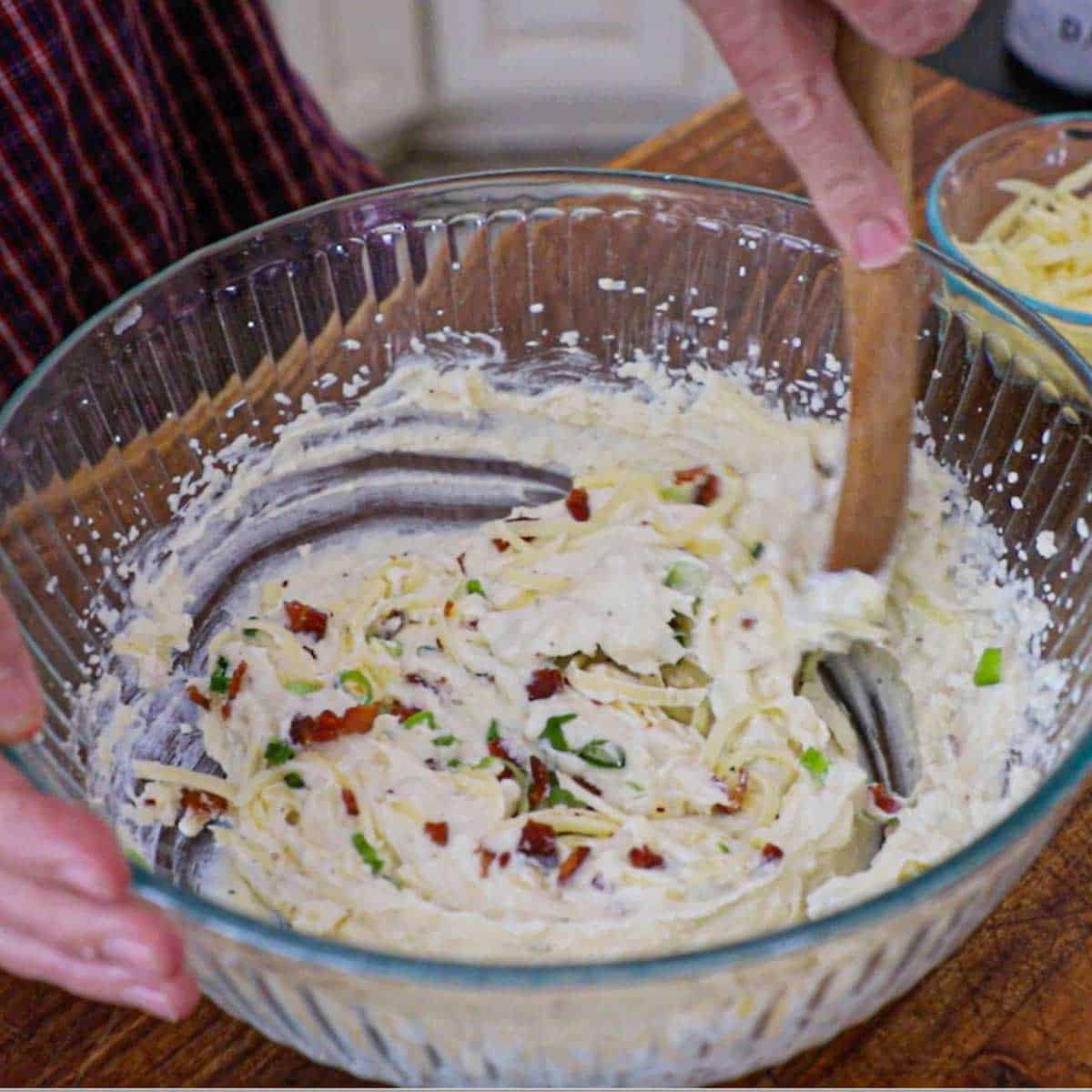 A person using a wooden spoon to stir in a large glass bowl whipped potatoes, herbed butter, sour cream, shredded white cheddar, crispy bacon bits, and chopped scallions.