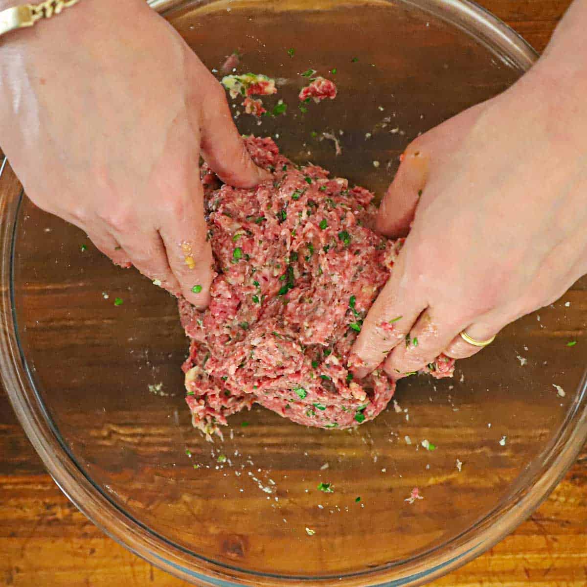 A person using his hands to combine the ingredients for Greek meatballs in a large glass bowl. 