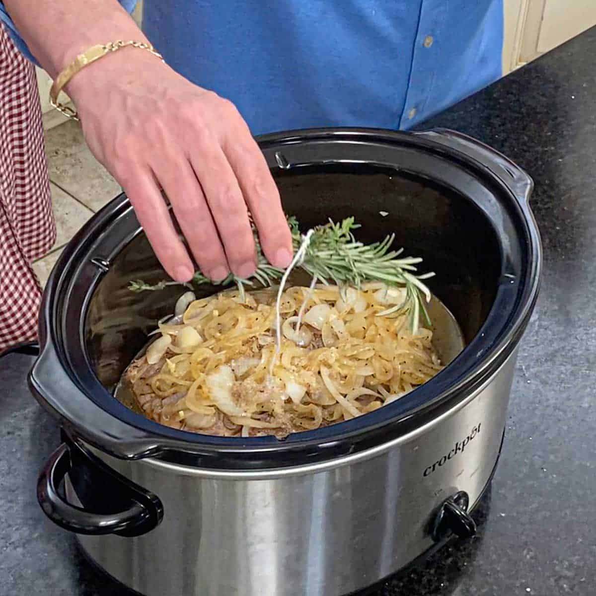 A person placing a bundle of fresh herbs that have been tied together with kitchen twine into a slow cooker filled with a partially seared chuck roast, beef stock, and sautéed onions.