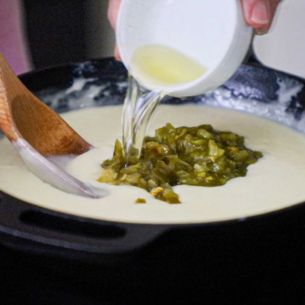 A person pouring jalapeño brine from a small white bowl into a skillet that is filled with white queso and topped with chopped pickled jalapeños.