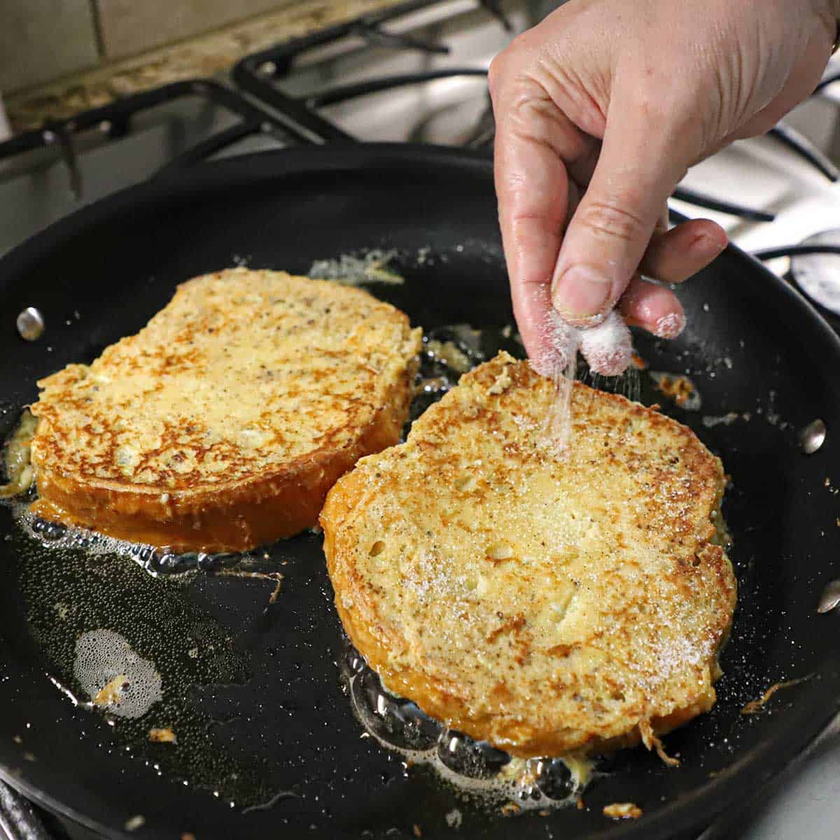 A person sprinkling superfine sugar over the surface of brioche French toast that are being lightly fried in a non-stick skillet on a gas stove.