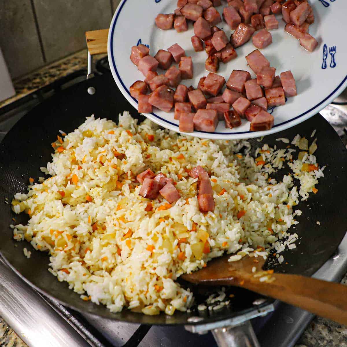A person transferring crispy chunks of ham from a white dinner plate into a large wok filled with fried rice, onions, carrots, and aromtics.