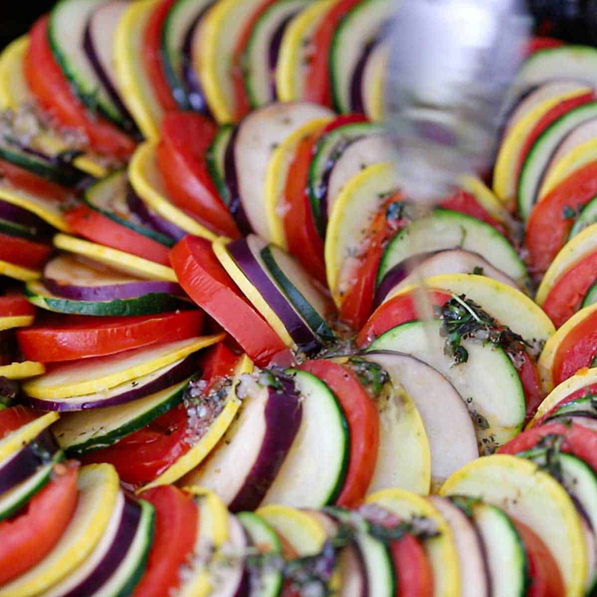 A person using a spoon to drizzle herbed oil over the top of an uncooked cast-iron skillet ratatouille.
