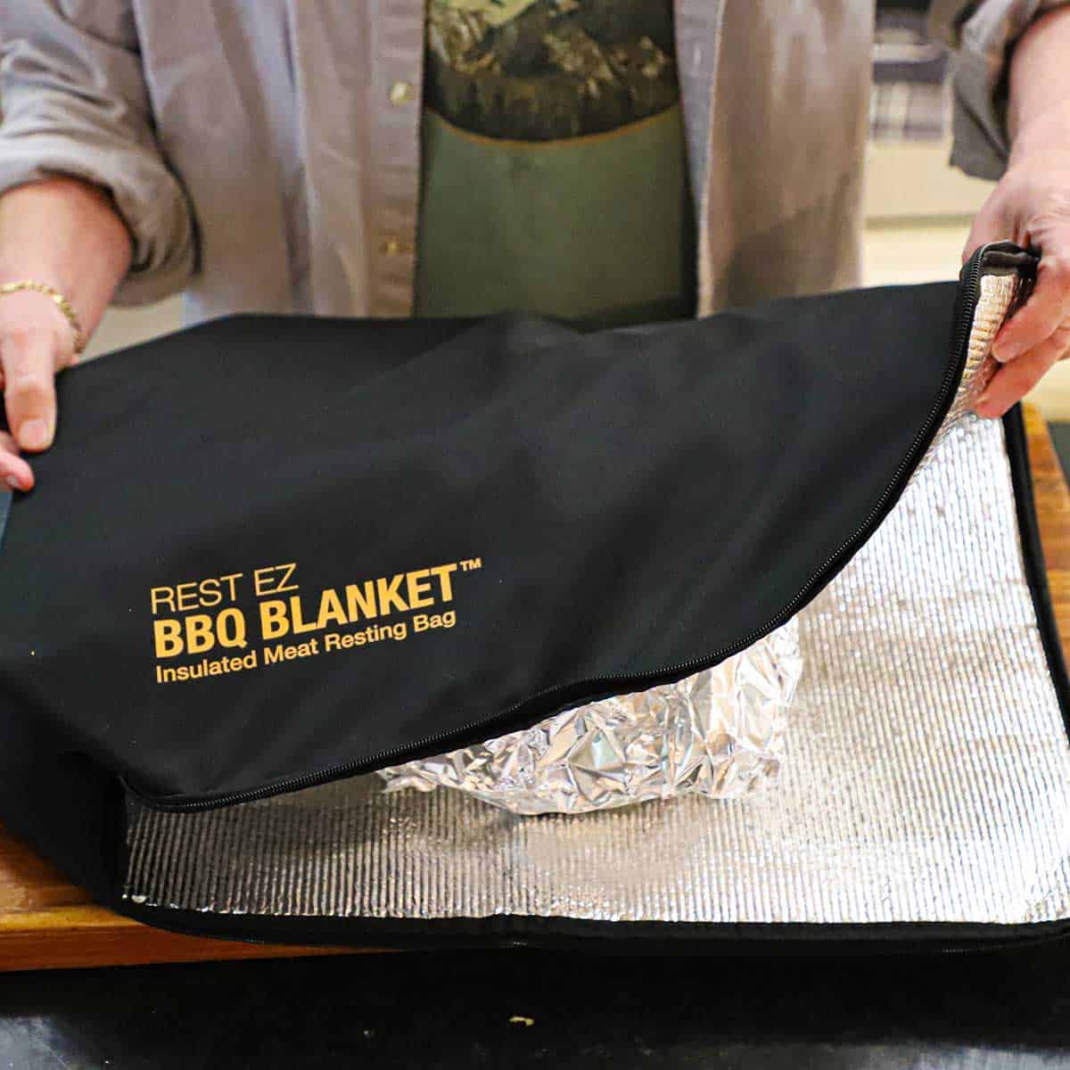 A person holding a BBQ resting blanket with a foil-wrapped smoked pork butt sitting inside the blanket.