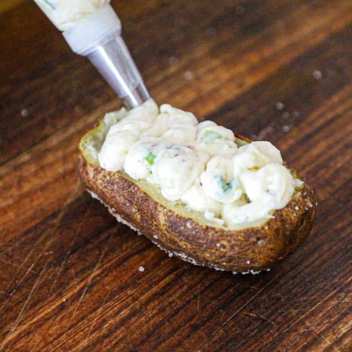 A person using a piping bag to pie softened twice potato filling into a shelled-out baked potato boat on a wooden cutting board.