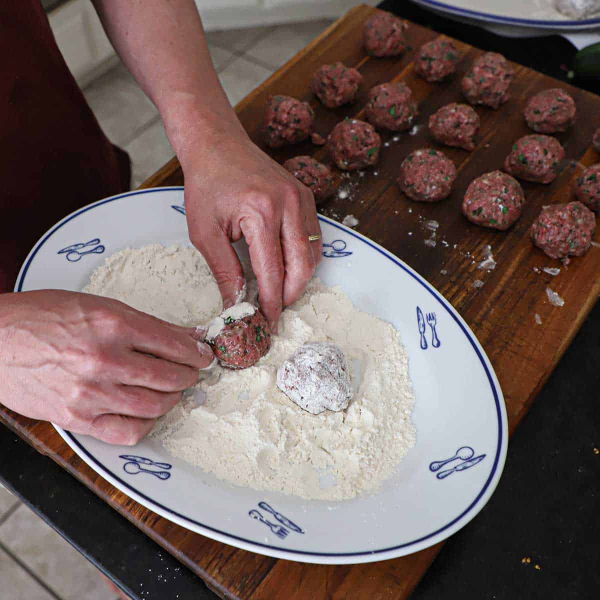 A person rolling uncooked Greek meatballs in flour on a large oval platter. 