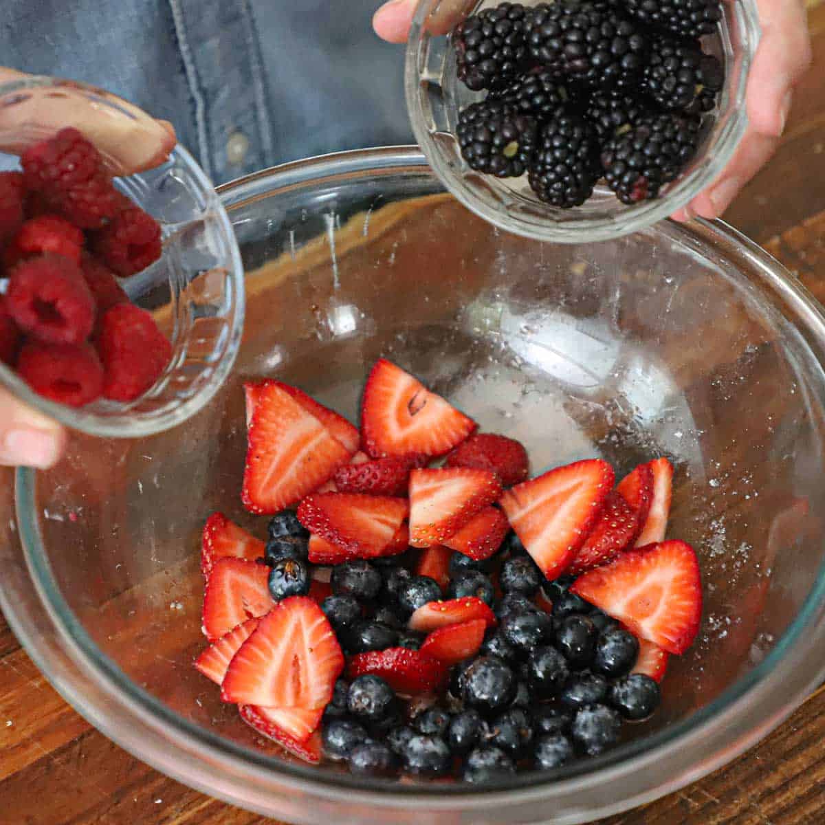A person holding a small bowl of blackberries in one hand and a small bowl of raspberries with his other hand both over a large bowl filled with macerated strawberries and blueberries.