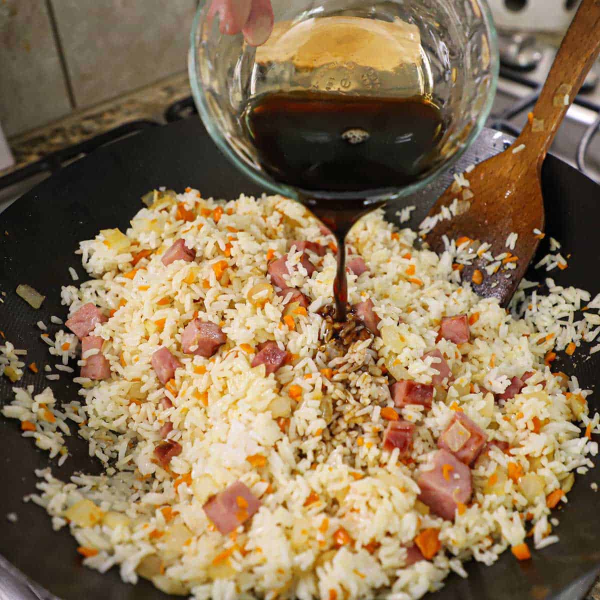 A person pouring a brown stir-fry sauce over leftover ham fried rice in a large wok on a gas stove.