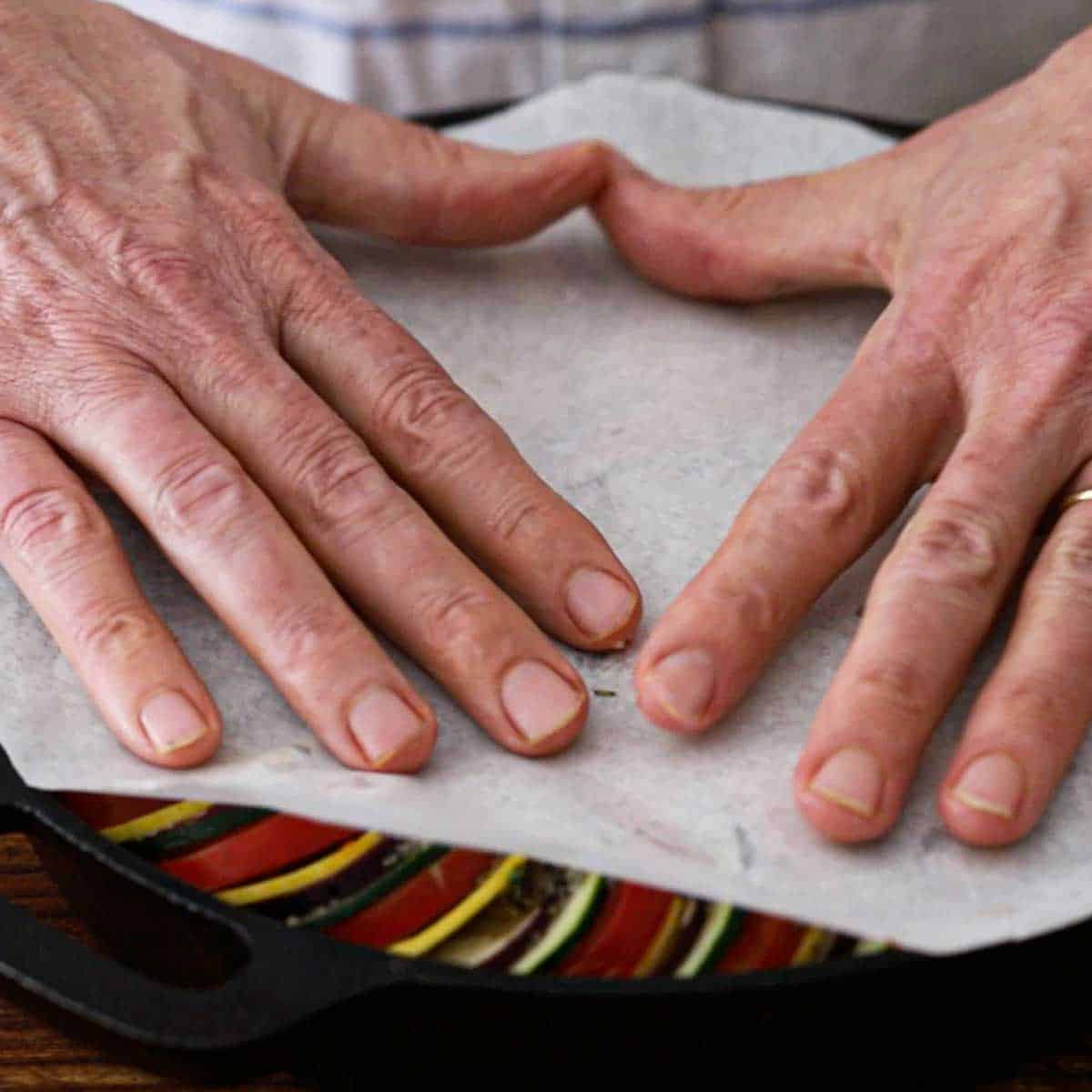 A person placing a circular piece of parchment paper over the top of an uncooked cast-iron skillet ratatouille.