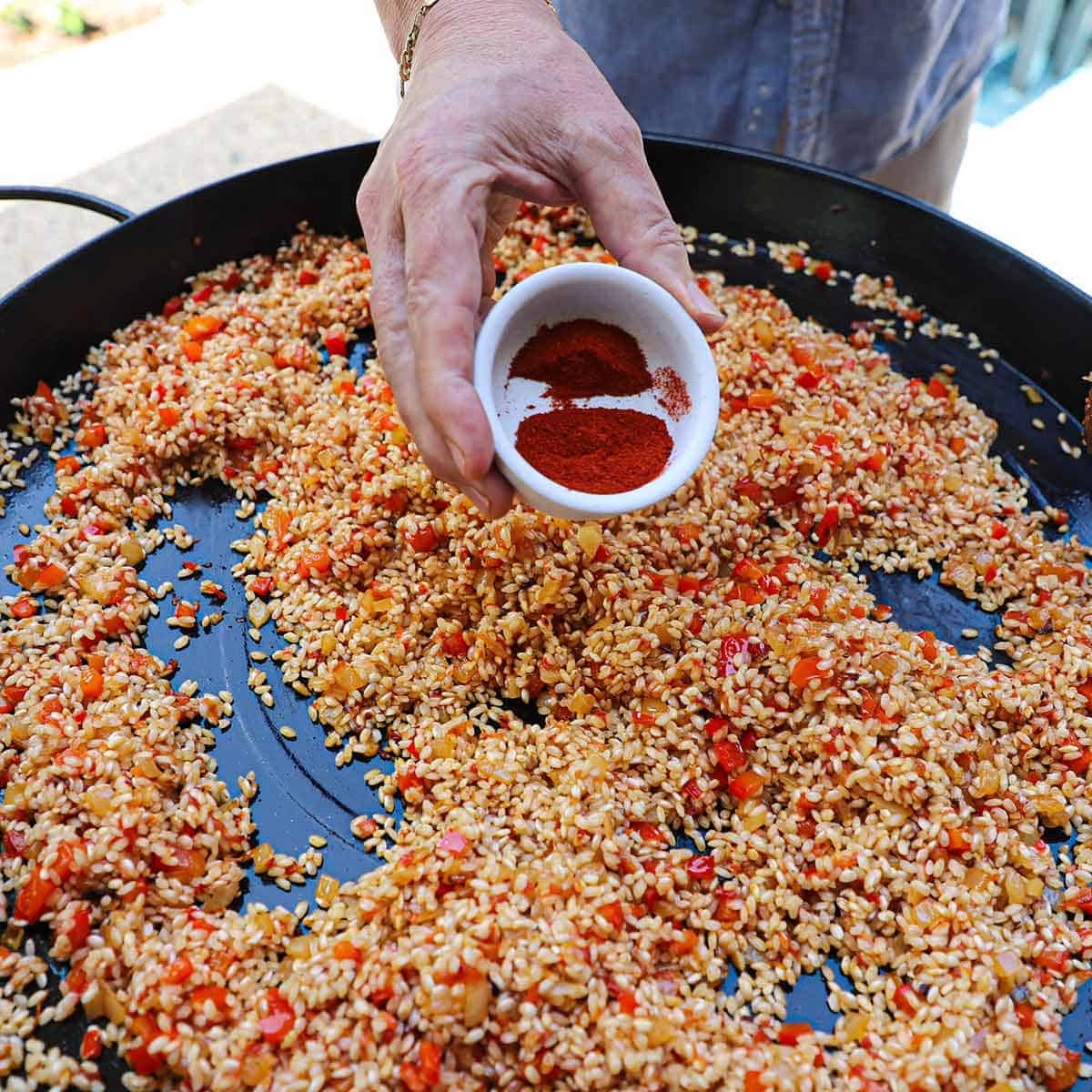 A person holding a small bowl of Spanish paprika over a large paella pan filled with toasted short-grain rice with sofrito.