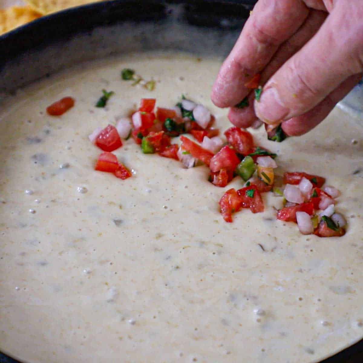 A person garnishing white queso blanco that is in a black cast-iron skillet with pico de gallo and chopped fresh cilantro.