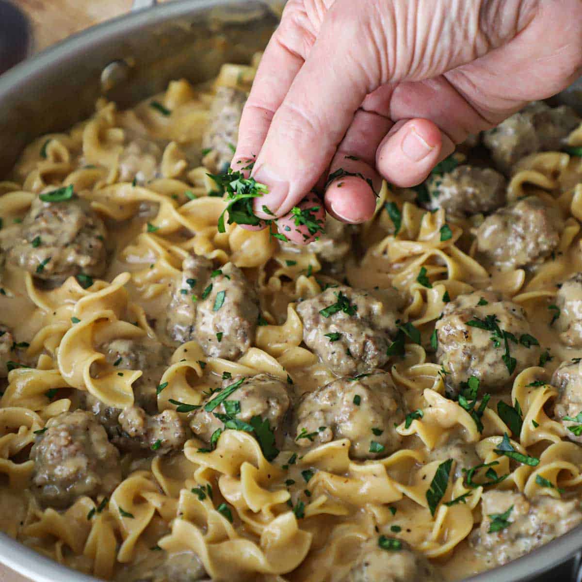 A person sprinkling chopped Italian parsley over a skillet filled with Swedish Meatball Pasta.