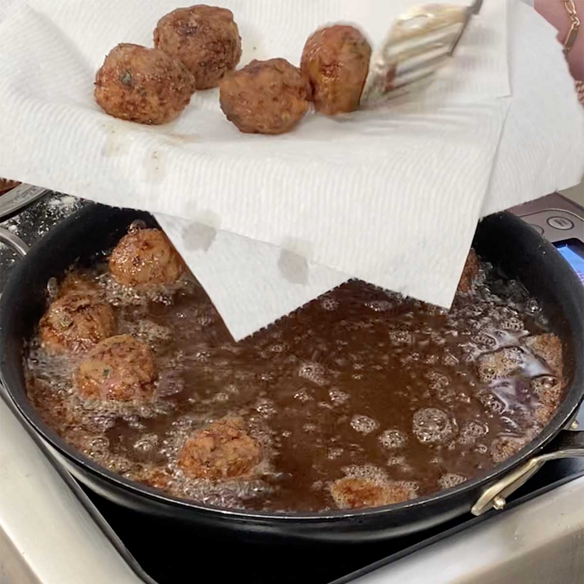 A person using a metal slotted spoon to place fried Greek meatballs from a skillet filled with hot oil onto a paper towel-line plate being held above the skillet.