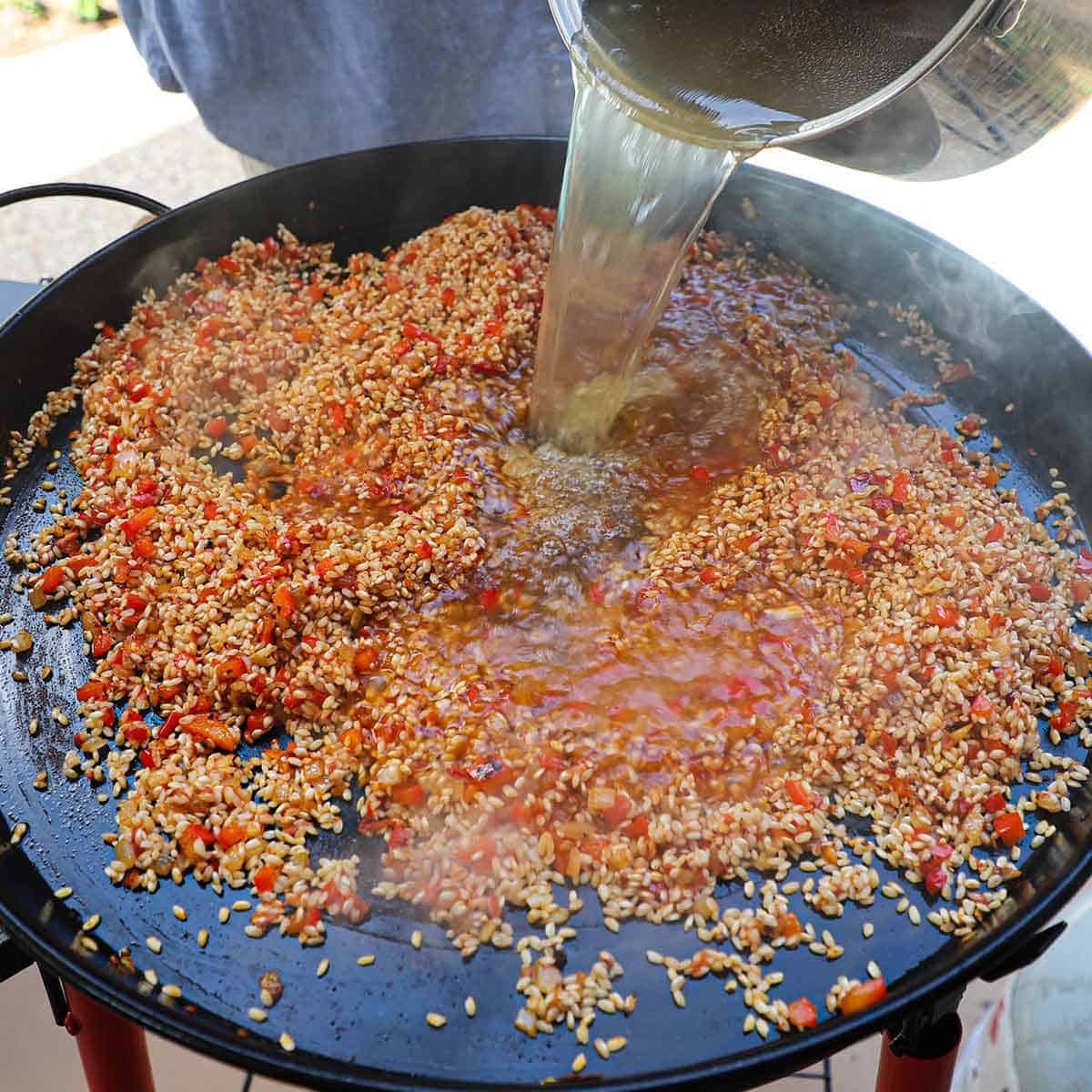 A person pouring hot shellfish stock from a stock pot into a large paella pan filled with toasted bomba rice mixed with sofrito.