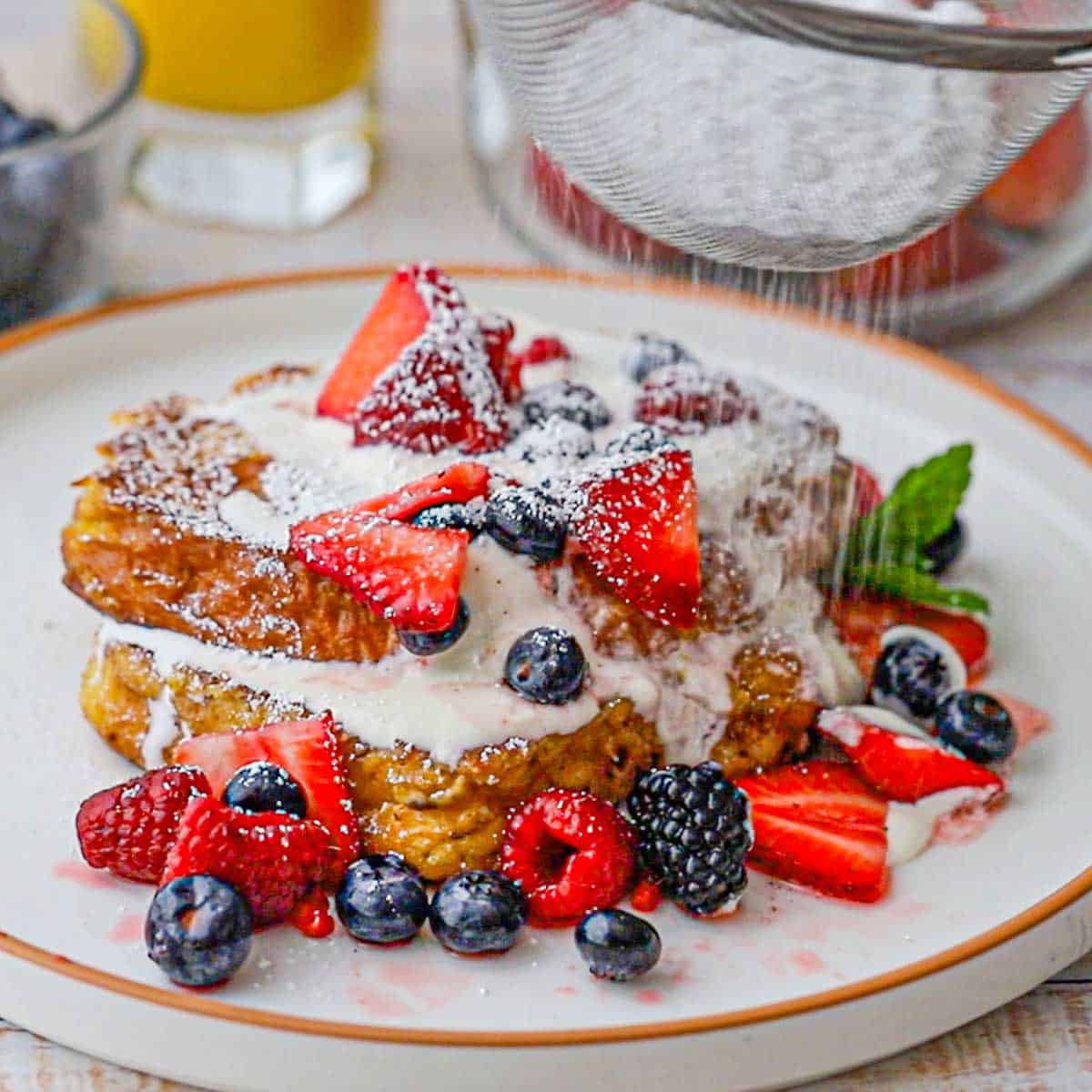 A person using a fine-mesh sieve to dust powdered sugar over the top of plate filled with caramelized brioche French toast topped with macerated berries and strawberries.