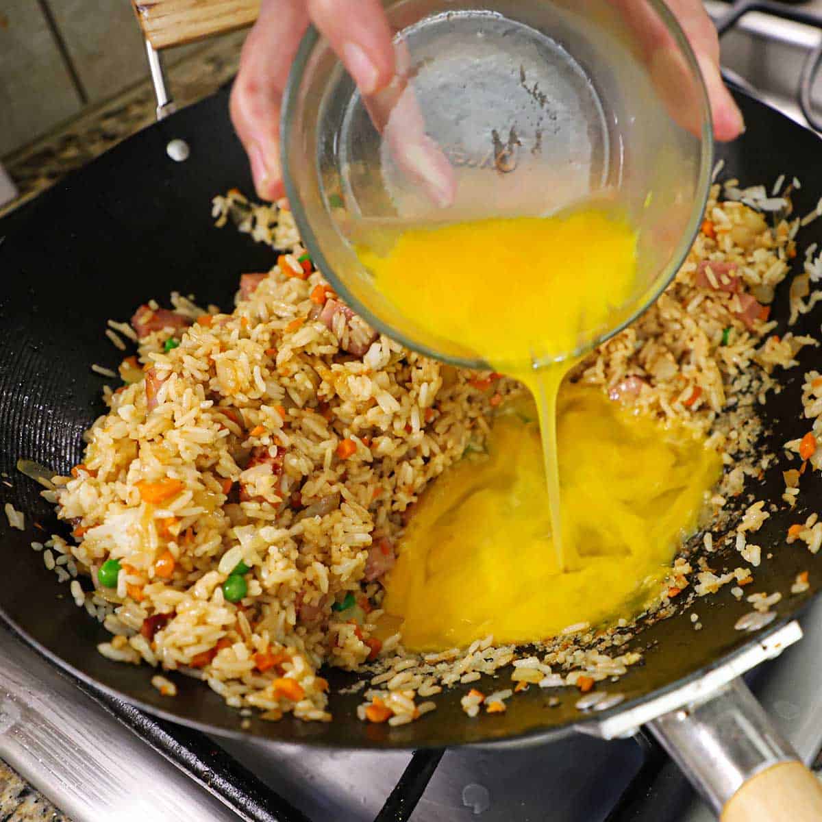 A person pouring three lightly beaten eggs from a small glass bowl into a wok that has ham fried rice pushed to the side, leaving space for the eggs to cook.