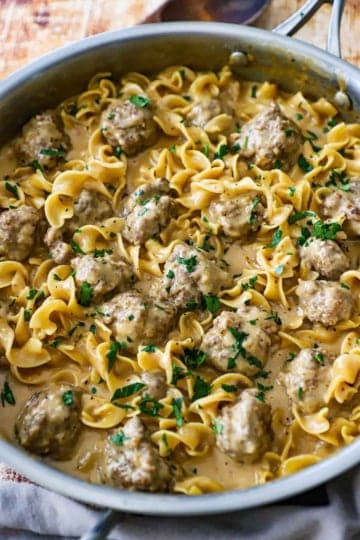 A large stainless steel skillet filled with Swedish meatball pasta that has been garnished with chopped Italian parsley.