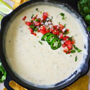 An overhead view of restaurant-style white queso blanco in a cast-iron skillet that has been garnished with pico de gallo, sliced jalapeño peppers, and chopped cilantro.