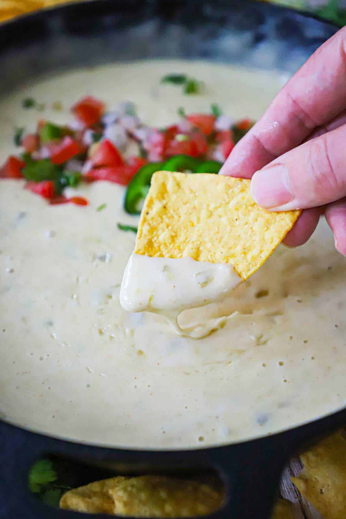 A person holding a triangular corn tortilla chip that he just dipped into a skillet filled with white queso blanco.