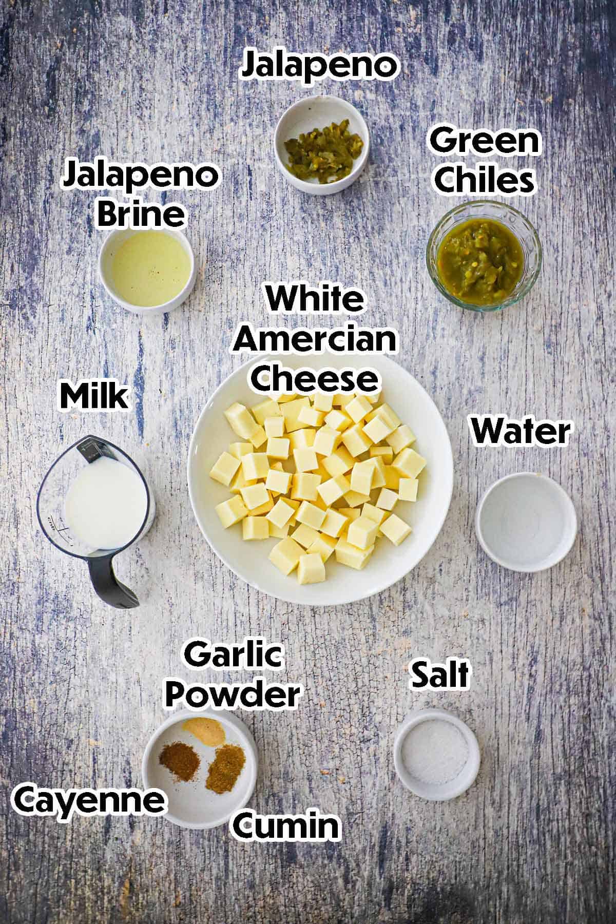 An arrangement of ingredients for white queso blanco on a grey wooden background including cube white American cheese, jalapeños, green chiles, jalapeño brine, milk, water, and seasonings.