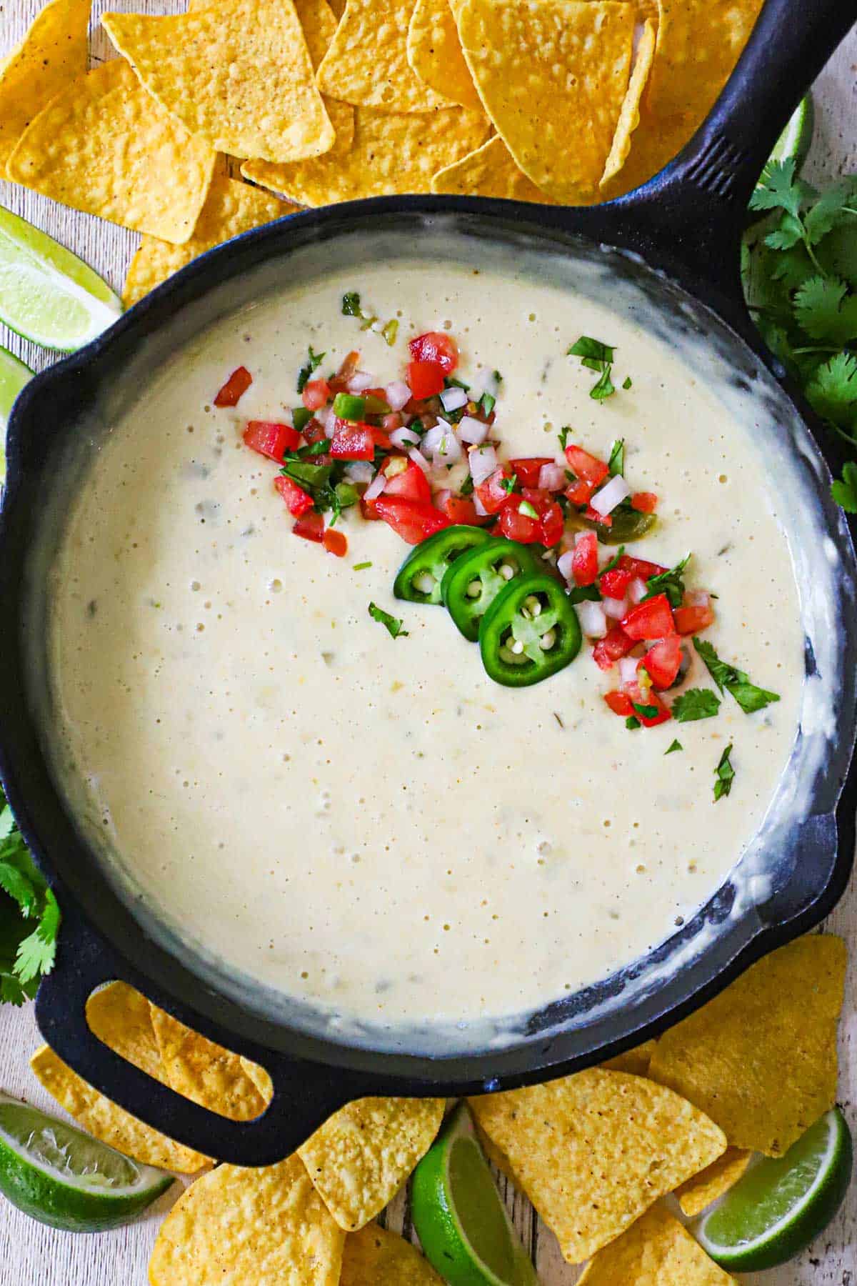 An overhead view of a cast-iron skillet filled with restaurant-style white queso (blanco) that has been garnished with pico de gallo, sliced jalapeños, and chopped cilantro.