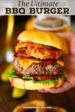 A person holding a massive BBQ Bacon burger topped with onion rings in front of two glasses of beer.