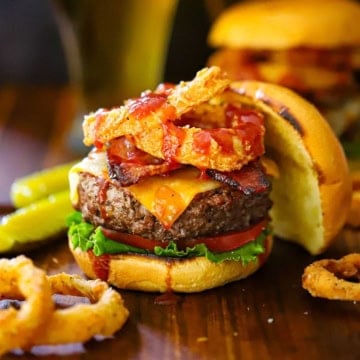 A close-up view of a BBQ Bacon Burger that is topped with crispy bacon, onion rings and drizzled with BBQ Sauce with more onion rings around the base of the burger.