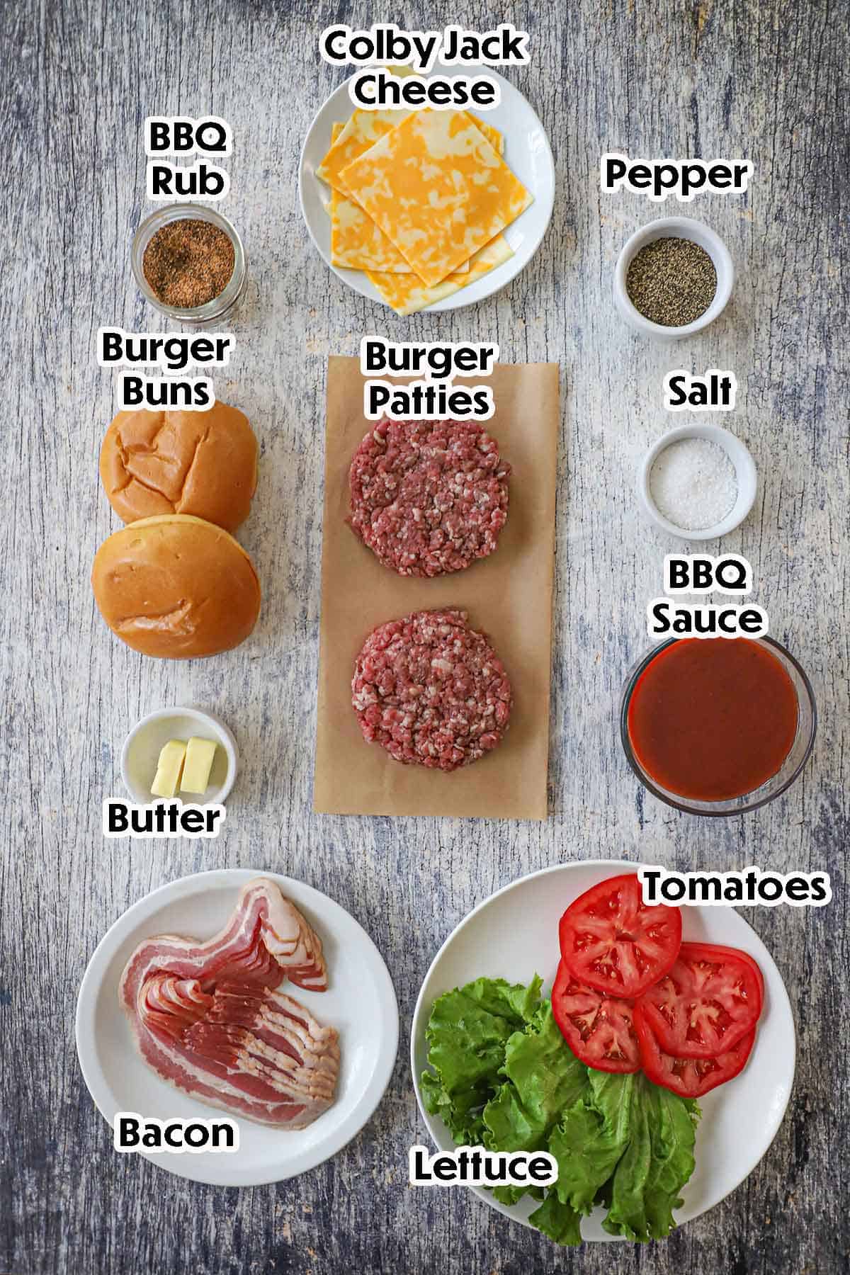An arrangement of ingredients for BBQ Bacon Burger on a grey wooden background including burger patties, BBQ sauce, bacon, cheese slices, buns, lettuce, tomaotes, and seasonings.