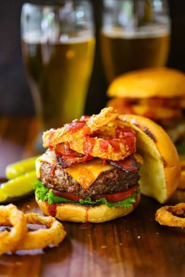 A straight-on view of a BBQ Bacon Burger that is topped with crispy bacon, onion rings and drizzled with BBQ Sauce with more onion rings around the base of the burger.