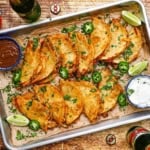 An overhead view of a large steel sheet pan filled with two rows of crispy baked chicken tacos with a bowl of enchilada sauce in one corner and another bowl of sour cream in the other corner.