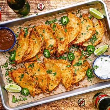 An overhead view of a large steel sheet pan filled with two rows of crispy baked chicken tacos with a bowl of enchilada sauce in one corner and another bowl of sour cream in the other corner.