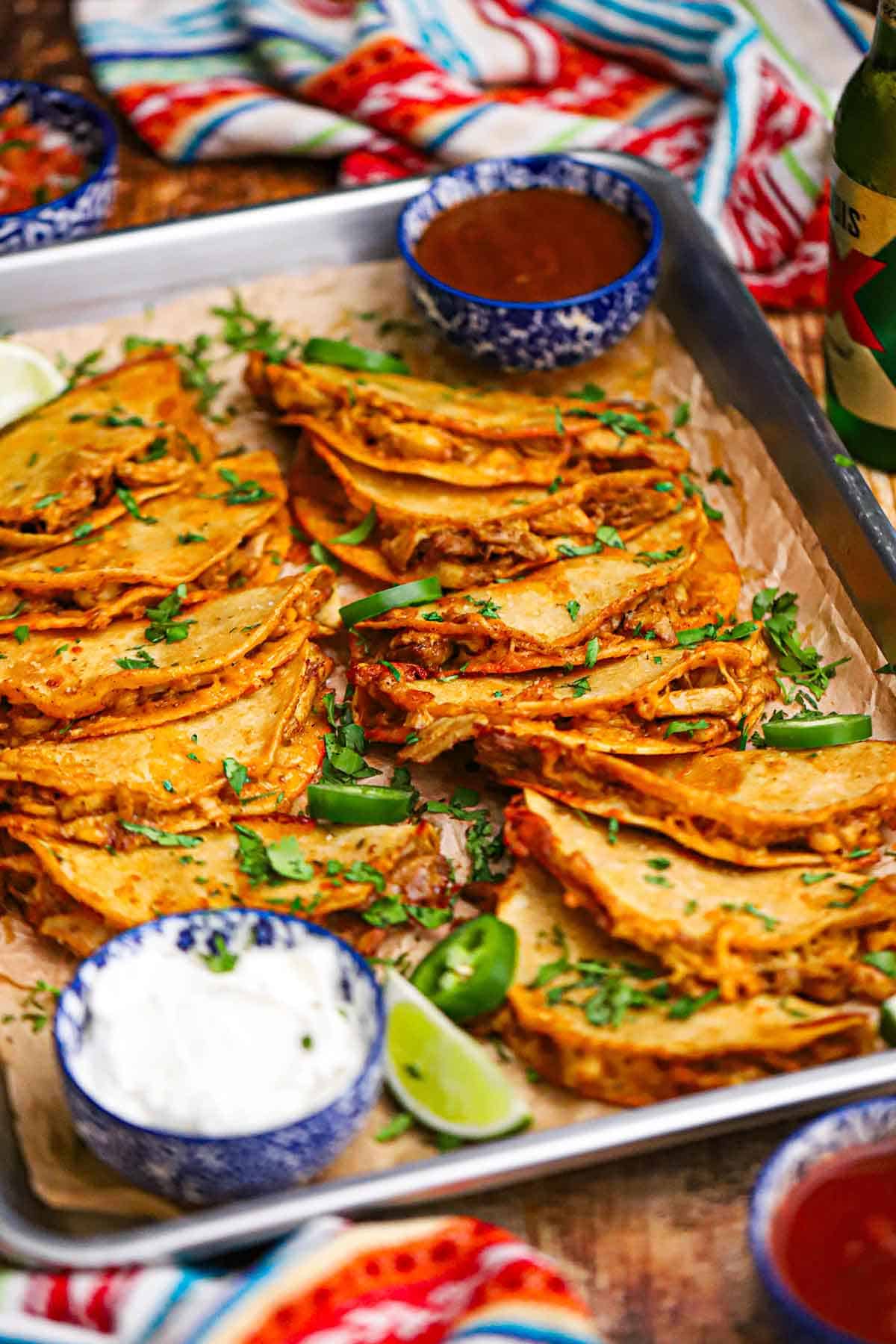 A large silver sheet pan filled with two rows of overlapping crispy baked chicken tacos garnished with chopped cilantro.
