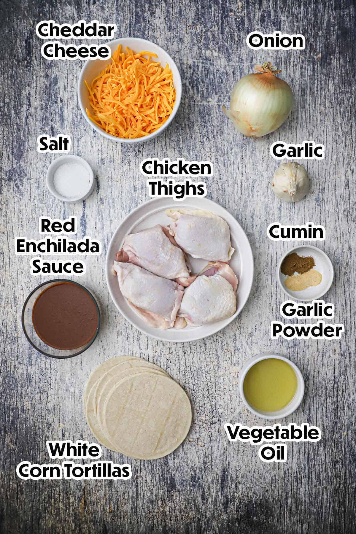 An arrangement of ingredients for Crispy Baked Chicken Tacos on a grey wooden background including chicken thighs, red enchilada sauce, cheddar cheese, corn tortillas, oil, garlic, onion, and seasonings.