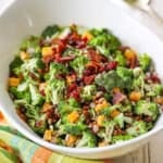 A close-up view of a white serving bowl filled with broccoli salad with bacon and topped with dried cranberries, cubed cheddar cheese, and sunflower seeds.