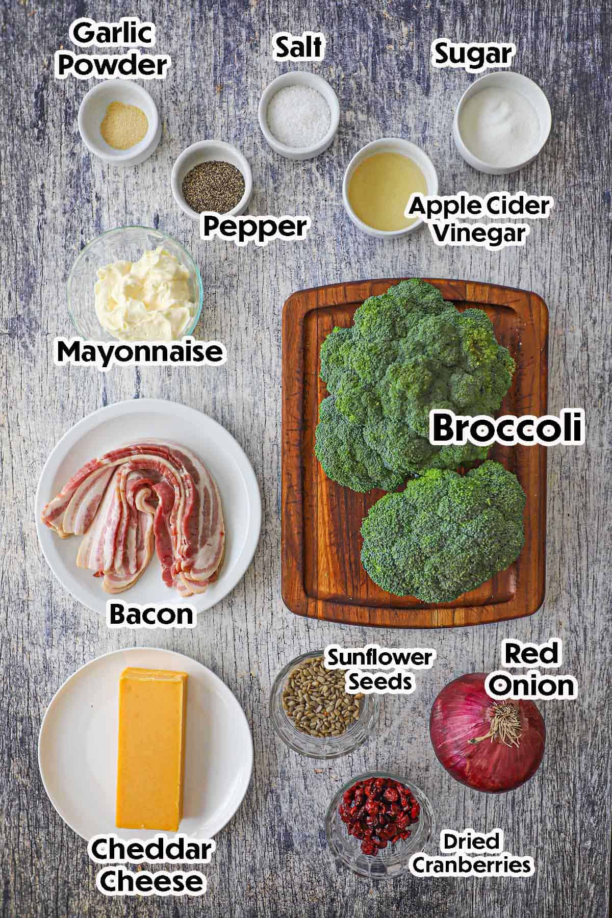An arrangement of ingredients for broccoli salad with bacon on a grey wooden background including two broccoli crowns, bacon, cheddar cheese, red onion, cranberries, sunflower seeds, bacon, mayonnaise, vinegar, and seasonings.