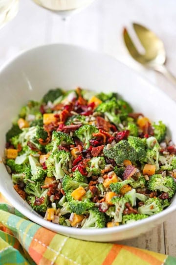 A close-up view of a white serving bowl filled with broccoli salad with bacon and topped with dried cranberries, cubed cheddar cheese, and sunflower seeds.