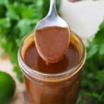 A person using a spoon to hold up a spoonful of easy homemade red enchilada sauce from a jar filled with the same.