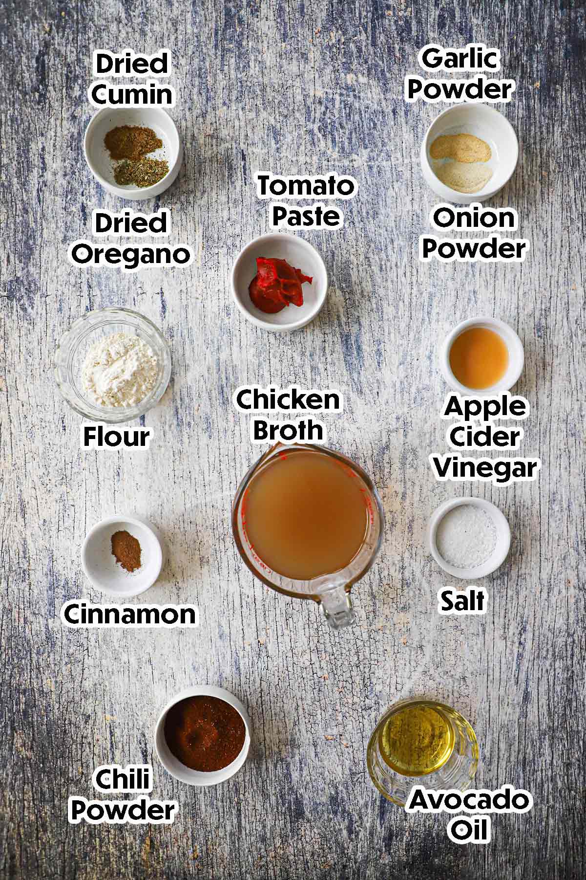 An arrangement of ingredients for Easy Homemade Red Enchilada Sauce on a grey wooden background including chicken broth, tomato paste, flour, oil, vinegar, and seasonings.