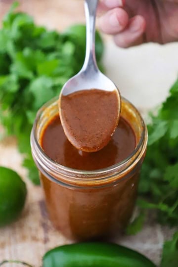 A person using a spoon to hold up a spoonful of easy homemade red enchilada sauce from a jar filled with the same.