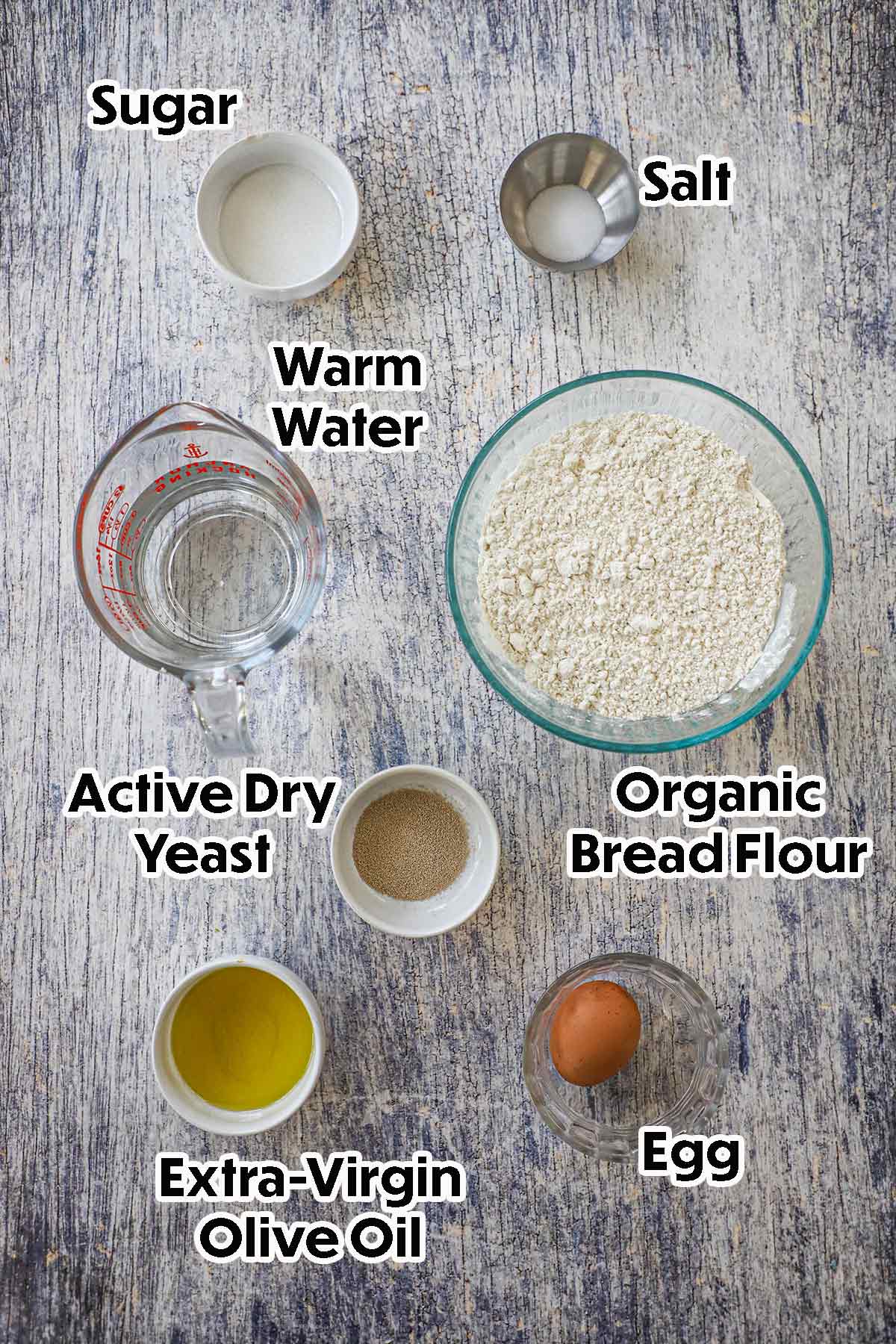An arrangement of ingredients for Homemade Italian Rolls on a grey wooden background including organic bread flour, active dry yeast, warm water, sugar, salt, olive oil, and an egg.