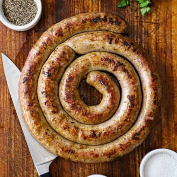 An overhead view of a circular rope of homemade Italian sausage the has been cooked and seared and is resting on a wooden cutting board.