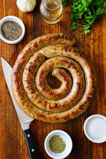 An overhead view of a circular rope of homemade Italian sausage the has been cooked and seared and is resting on a wooden cutting board and is surround by small bowls of seasonings.