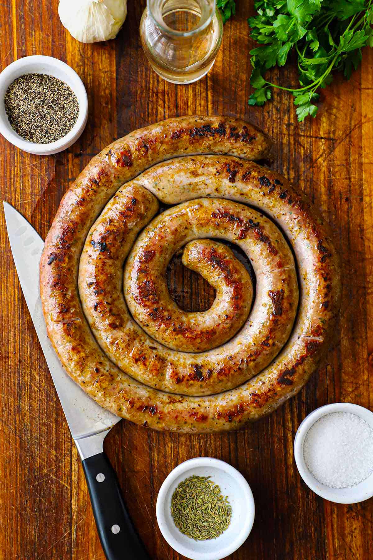 An overhead view of a circular rope of homemade Italian sausage the has been cooked and seared and is resting on a wooden cutting board and is surround by small bowls of seasonings.